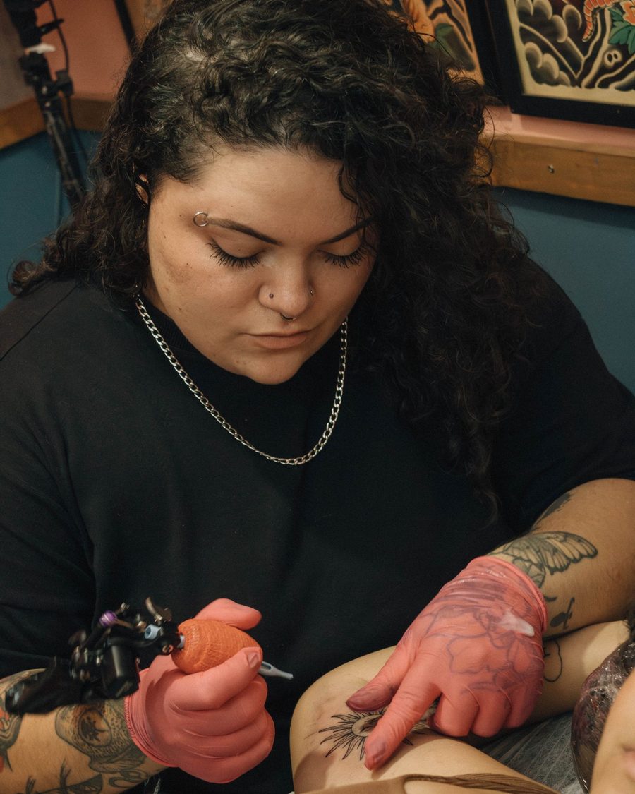 Rosa, a tattoo artist with dark curly hair, tattooing a client's arm in red gloves
