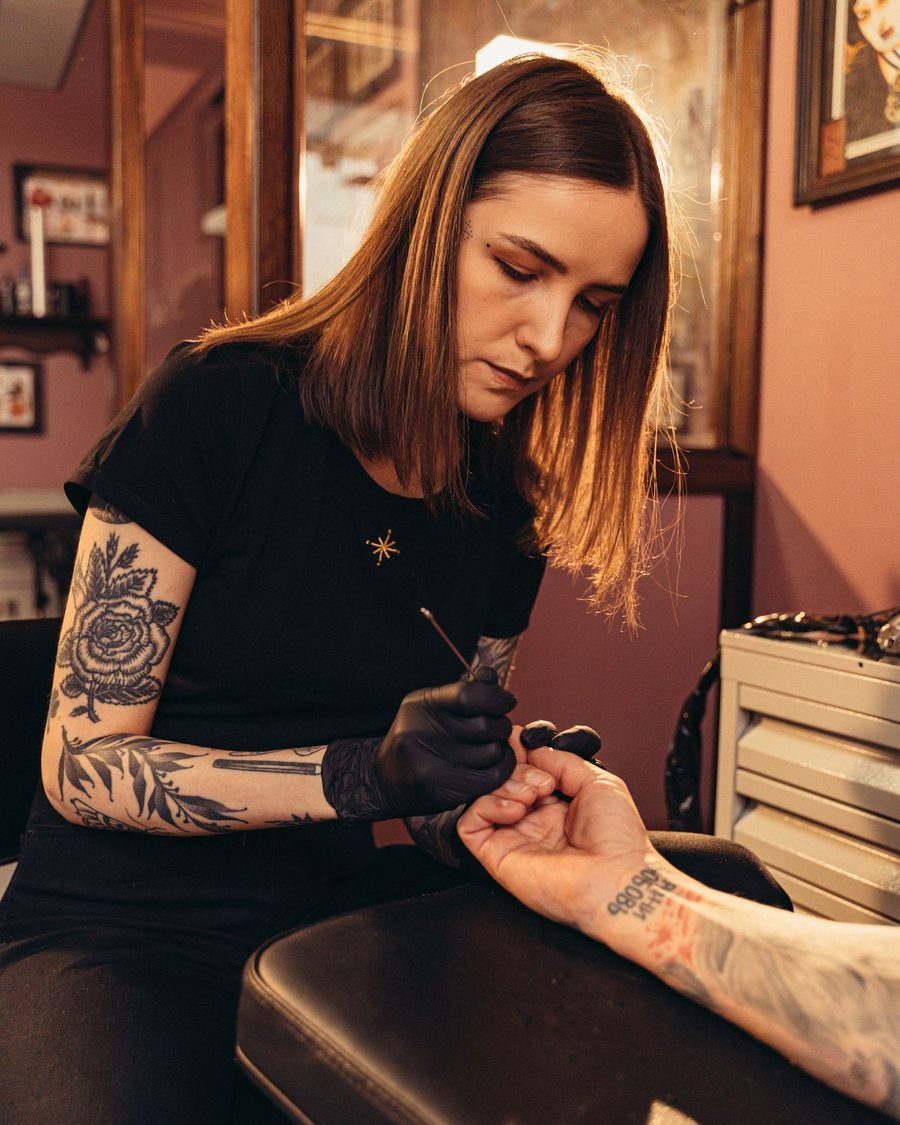 Wren, a tattoo artist with long hair, working on a client's wrist at a padded arm rest