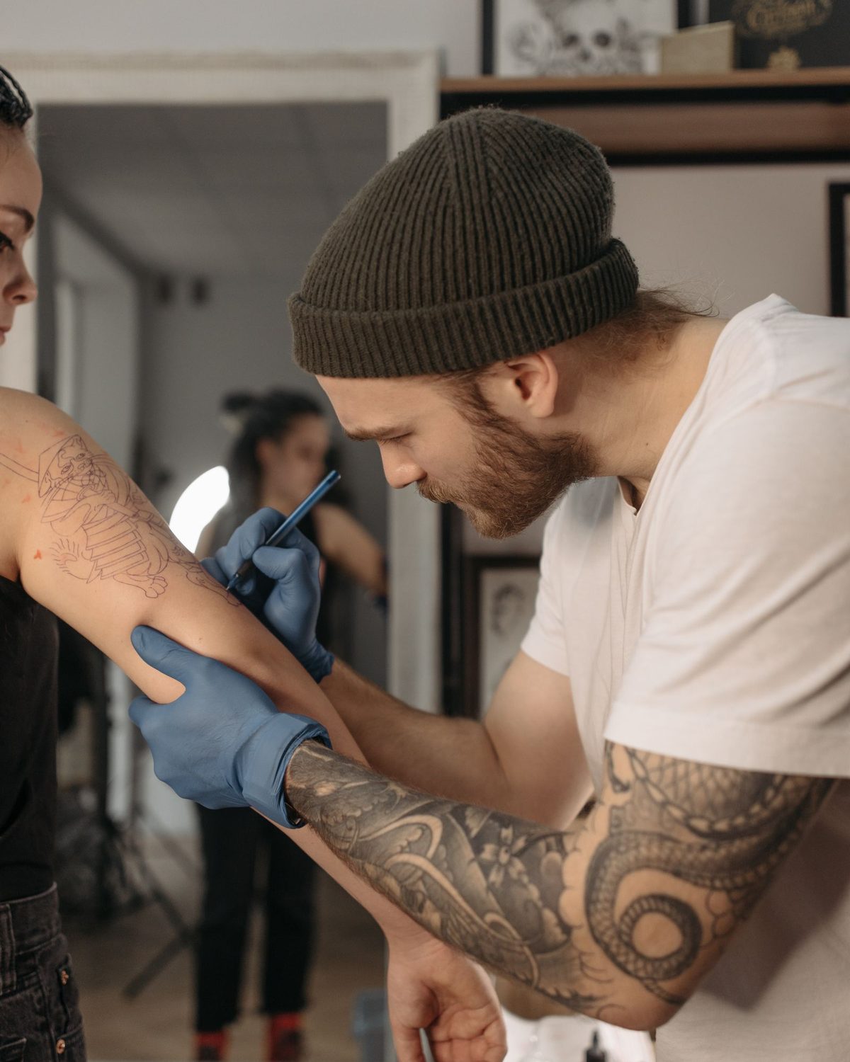 A tattoo artist in a beanie and blue gloves tattooing a client’s forearm in a bright studio