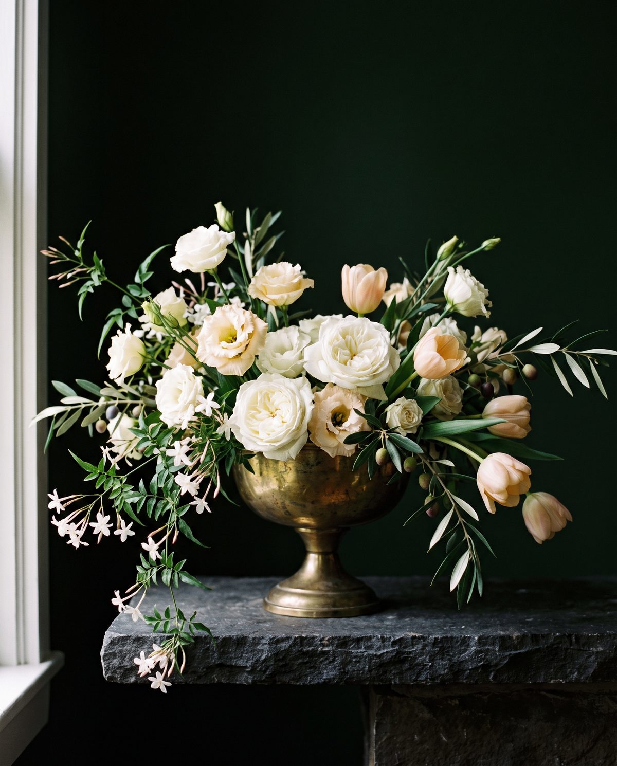 A low centerpiece of white roses, peach tulips, and trailing jasmine in a brass compote bowl.